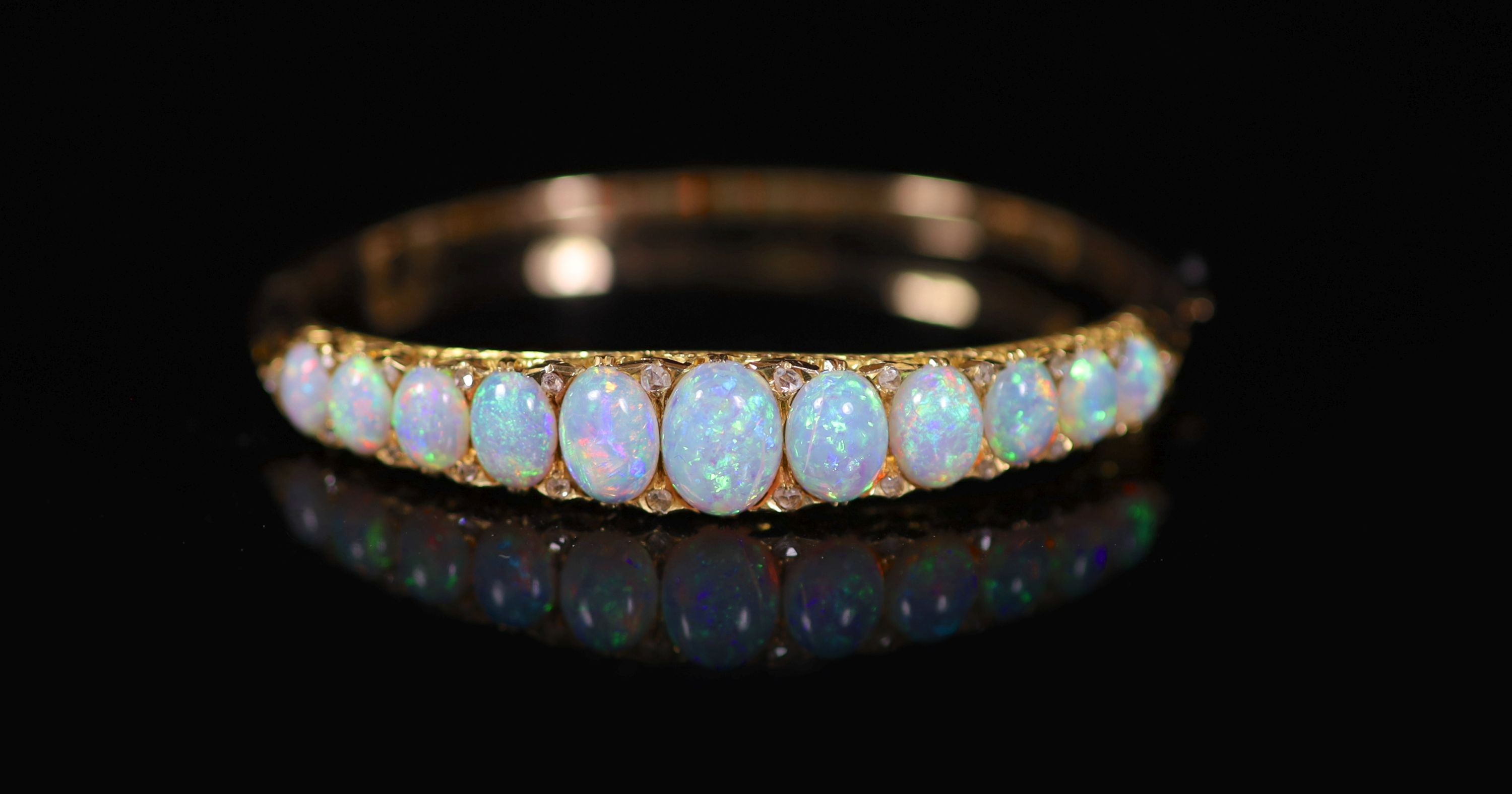 An Edwardian gold and graduated eleven stone white opal set hinged bracelet, with diamond chip spacers